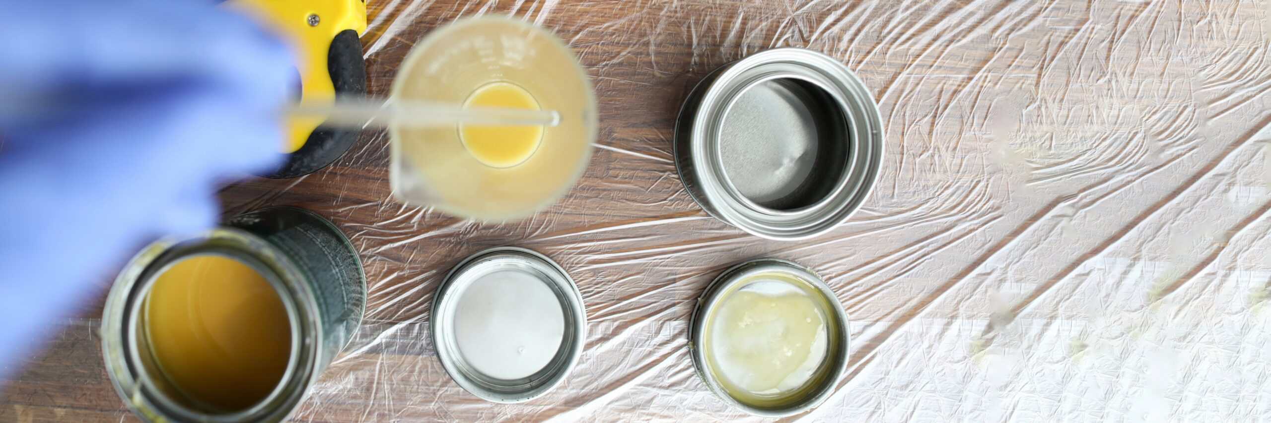 A gloved hand mixes yellow liquid in a beaker beside open metal containers with lids, all on a plastic-covered wooden surface.