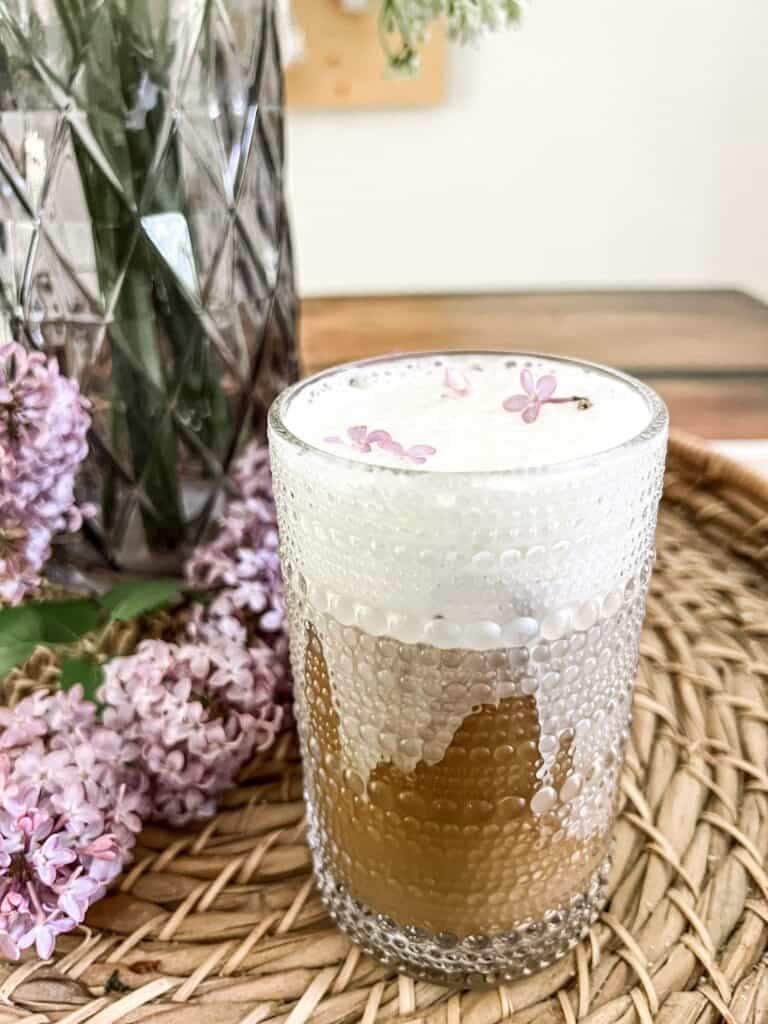 A glass of iced coffee with a thick layer of foam on top, garnished with small purple flowers, sits on a wicker tray next to lilac blossoms and a textured vase.