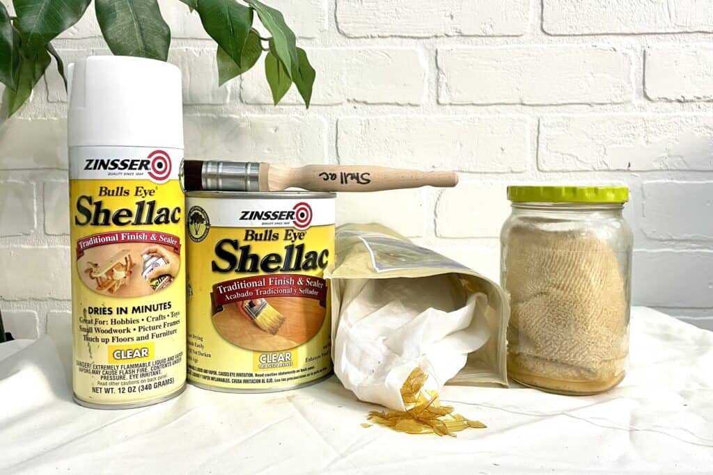A can and a spray can of Zinsser shellac, a paintbrush, a jar with a yellowish liquid, a bag of shellac flakes, and a piece of mesh are displayed on a white surface.