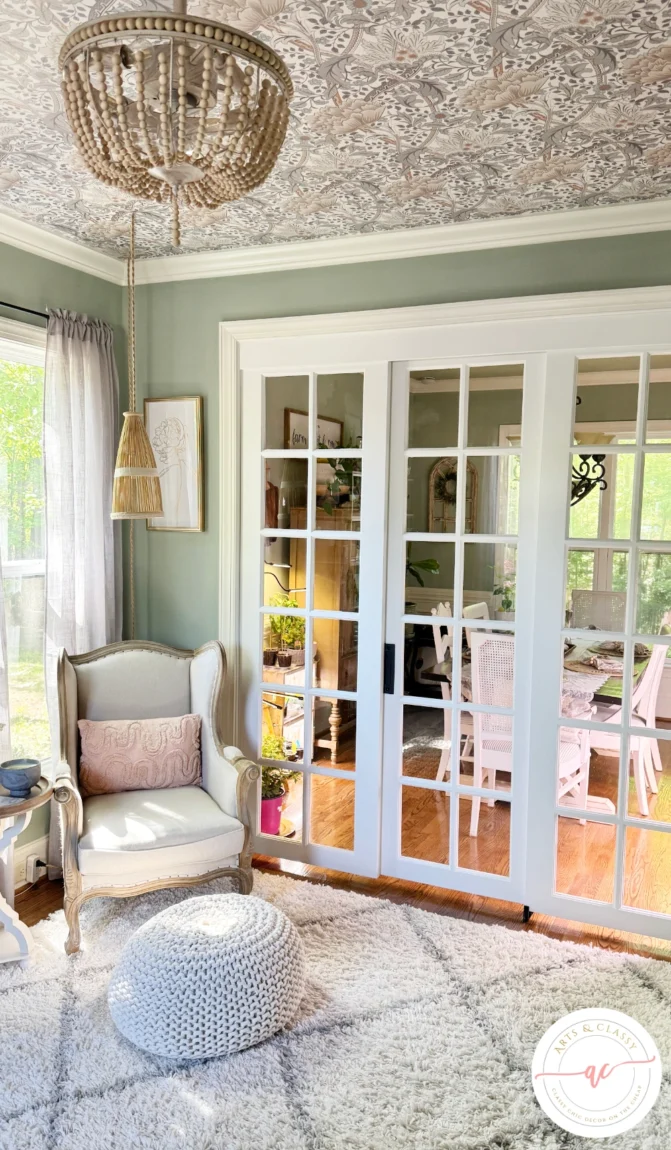 A cozy sitting area with a cushioned armchair, knitted pouf, and soft rug. French doors open to a sunlit room with plants and a dining table. Patterned ceiling and beaded chandelier overhead.