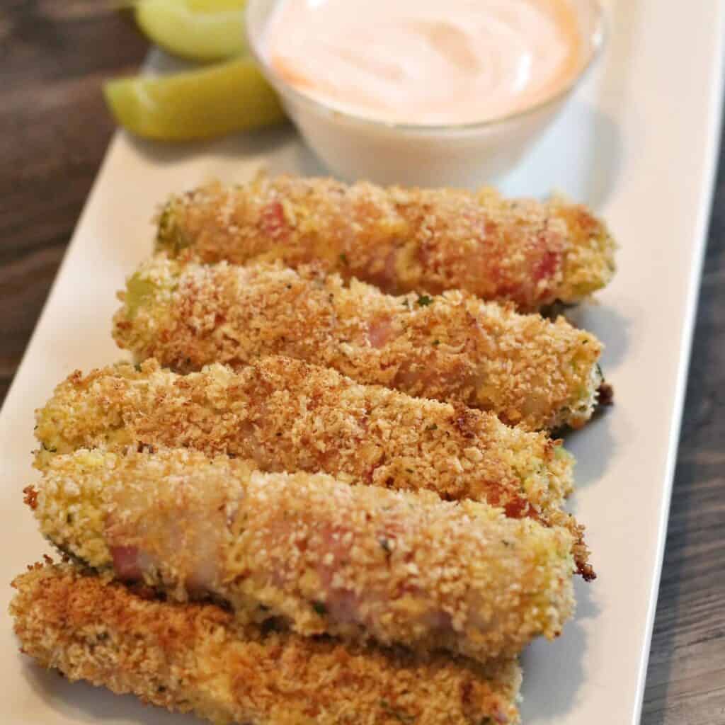 Five breaded and baked zucchini sticks are arranged on a white plate with a small bowl of creamy dipping sauce and two lemon wedges.