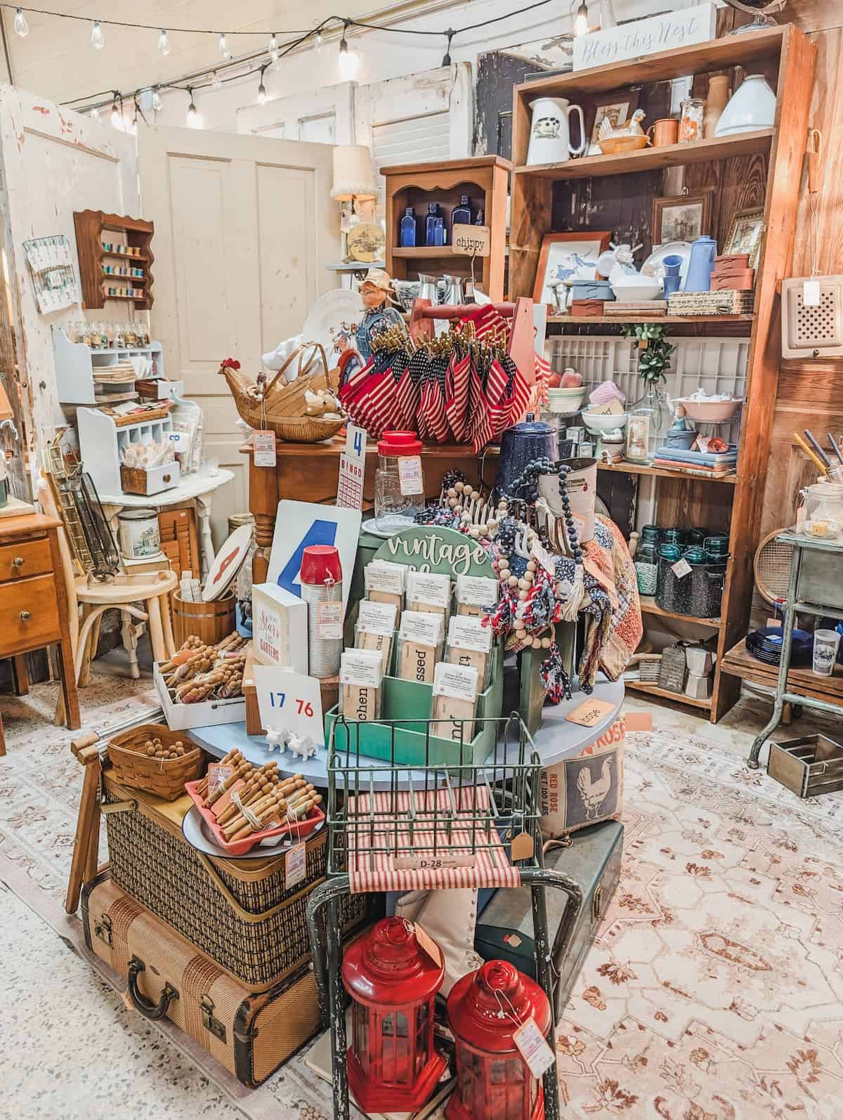 A vintage shop display with various home goods, including baskets, textiles, kitchenware, suitcases, red lanterns, and decorative items arranged on shelves and tables.