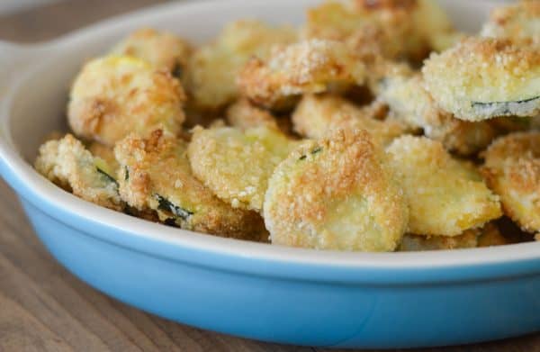 A blue dish filled with breaded and baked zucchini slices sits on a wooden surface.