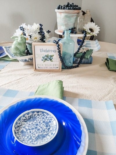 A table setting with blue and white plates, a green napkin, a green ceramic bird, a sign about blueberries, white flowers, and a pot of blueberries as the centerpiece.