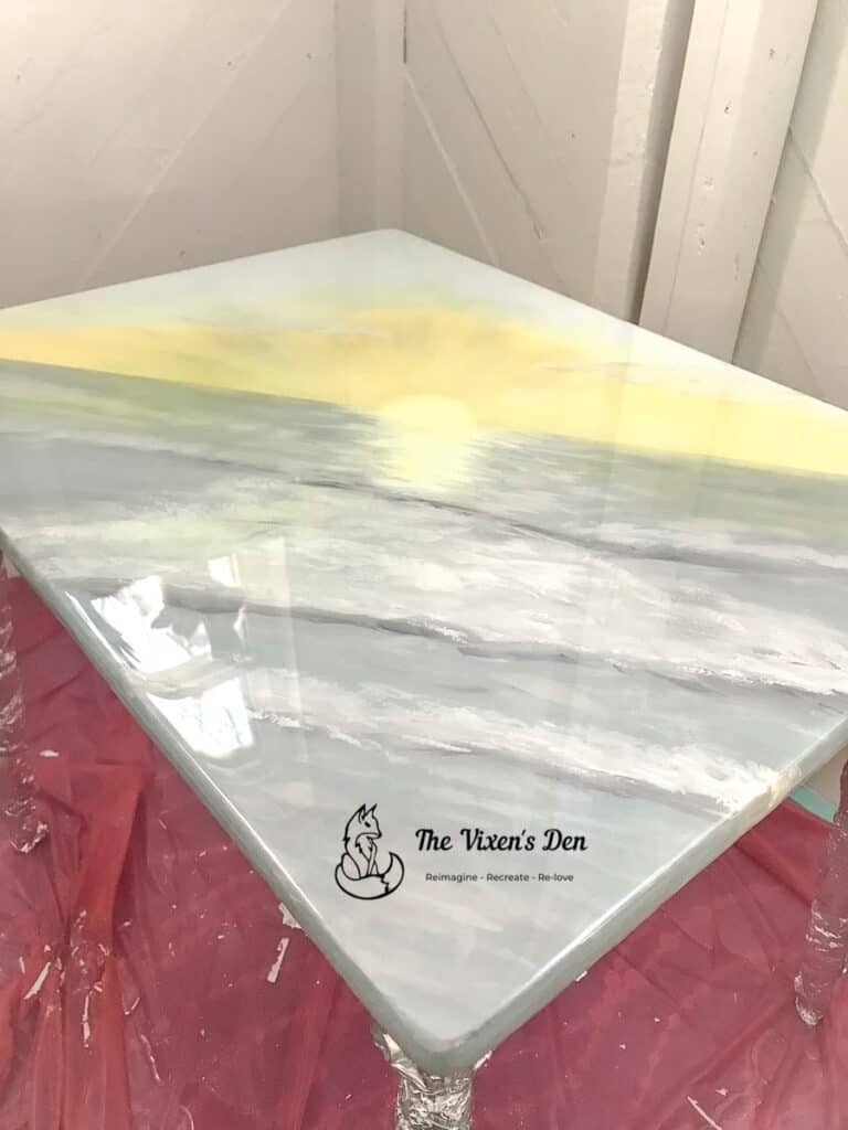 Square table with a glossy surface featuring a painted landscape of a yellow sun over water, with "The Vixen's Den" logo in one corner.