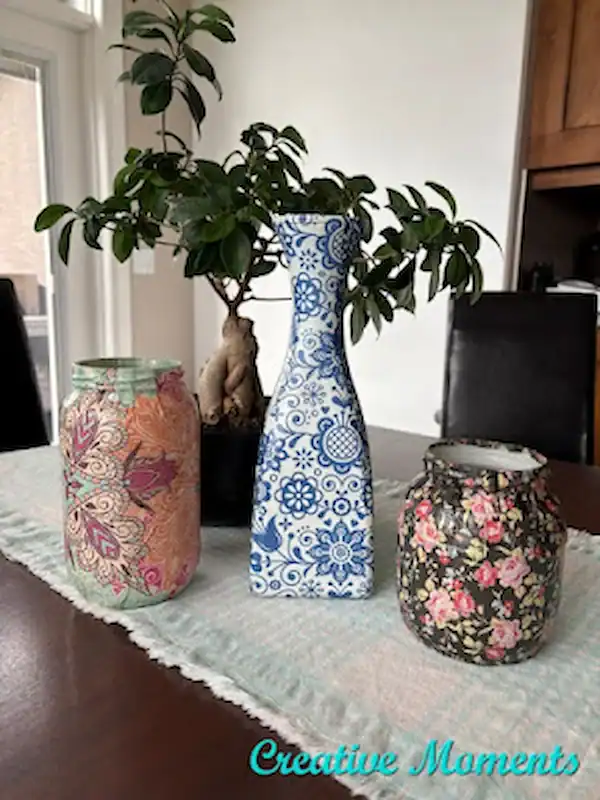 Three decorative jars with colorful patterns are displayed on a table runner with a potted plant and chairs in the background. The text “Creative Moments” appears in the corner.