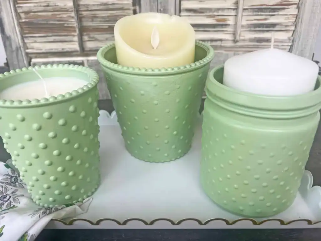 Three pastel green textured candle holders with white candles are arranged on a white tray in front of rustic wooden shutters.