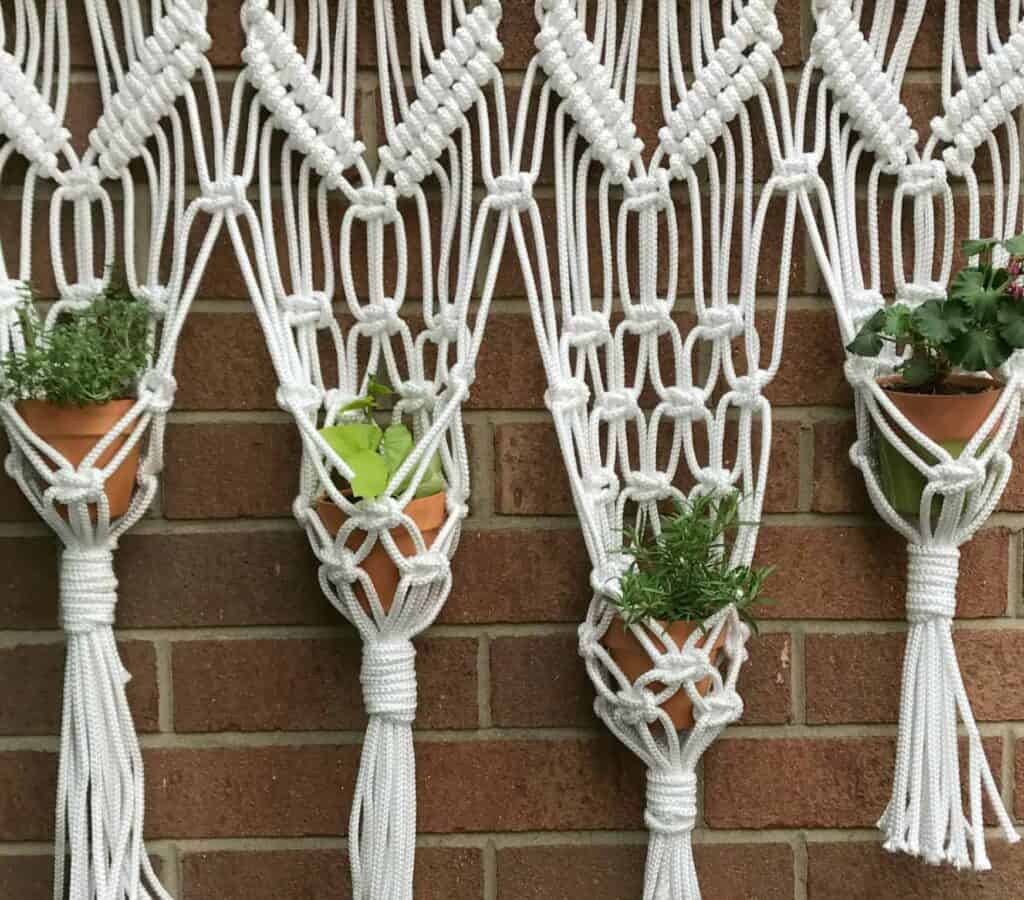 Macrame plant holders with small potted plants hang against a brick wall, each holder made of white rope in a geometric pattern.