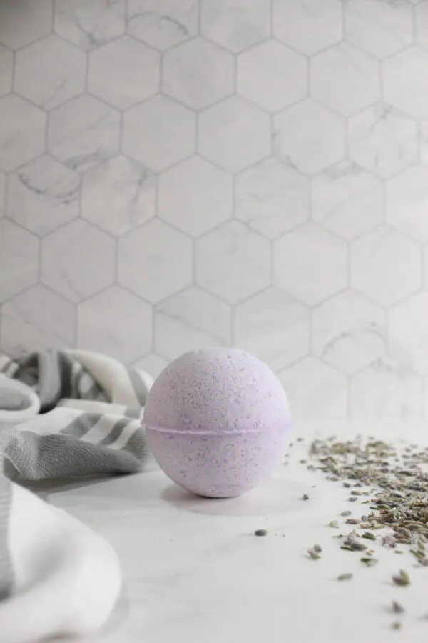 A round, light purple bath bomb sits on a white surface next to a gray-striped towel and scattered dried lavender buds, with a hexagon-tiled wall in the background.