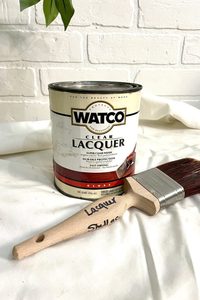 A can of Watco clear lacquer and a paintbrush labeled "Lacquer" and "Shellac" resting on a white fabric surface in front of a white brick wall.
