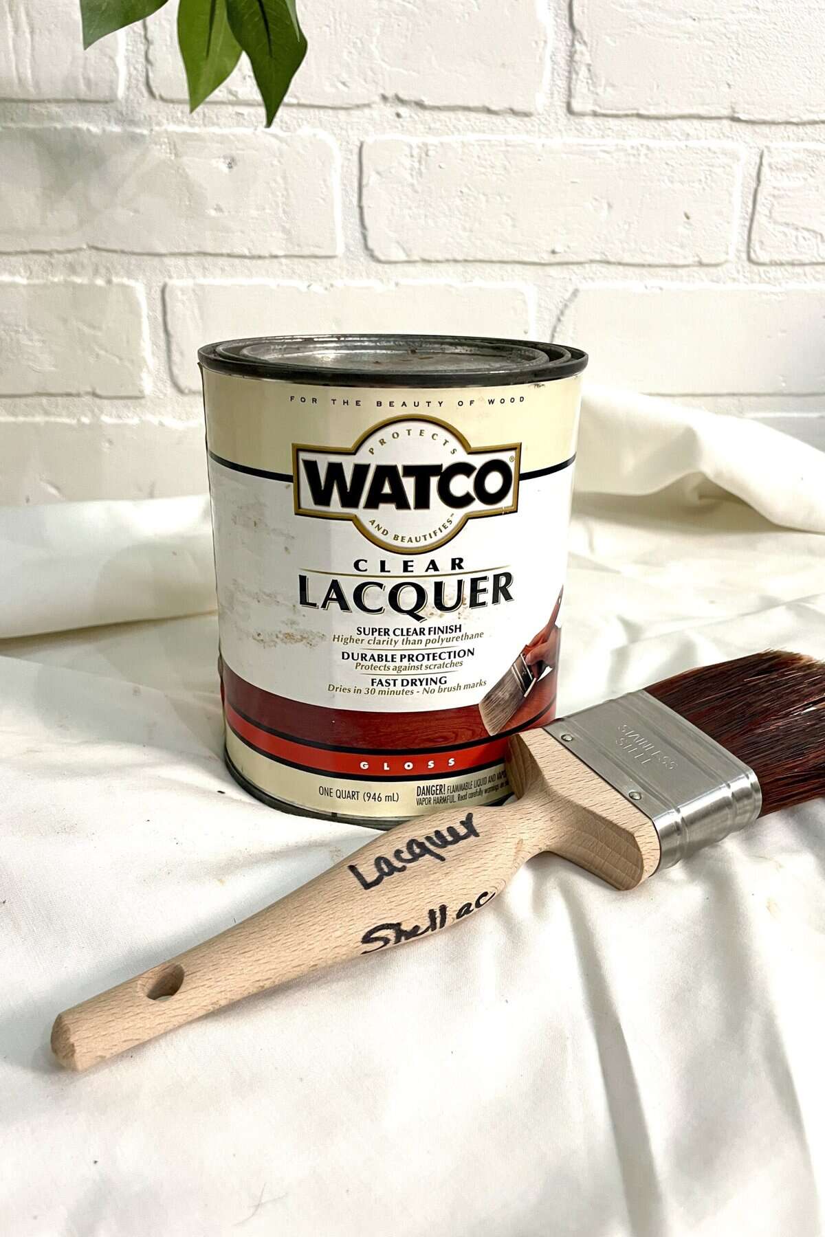 A can of Watco clear lacquer and a paintbrush labeled "Lacquer" and "Shellac" resting on a white fabric surface in front of a white brick wall.
