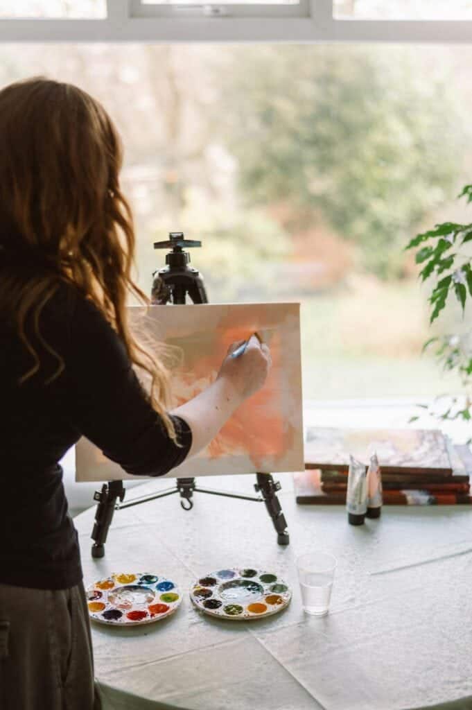 A person with long hair paints on a canvas set on an easel near a window, with paint palettes and brushes on the table.