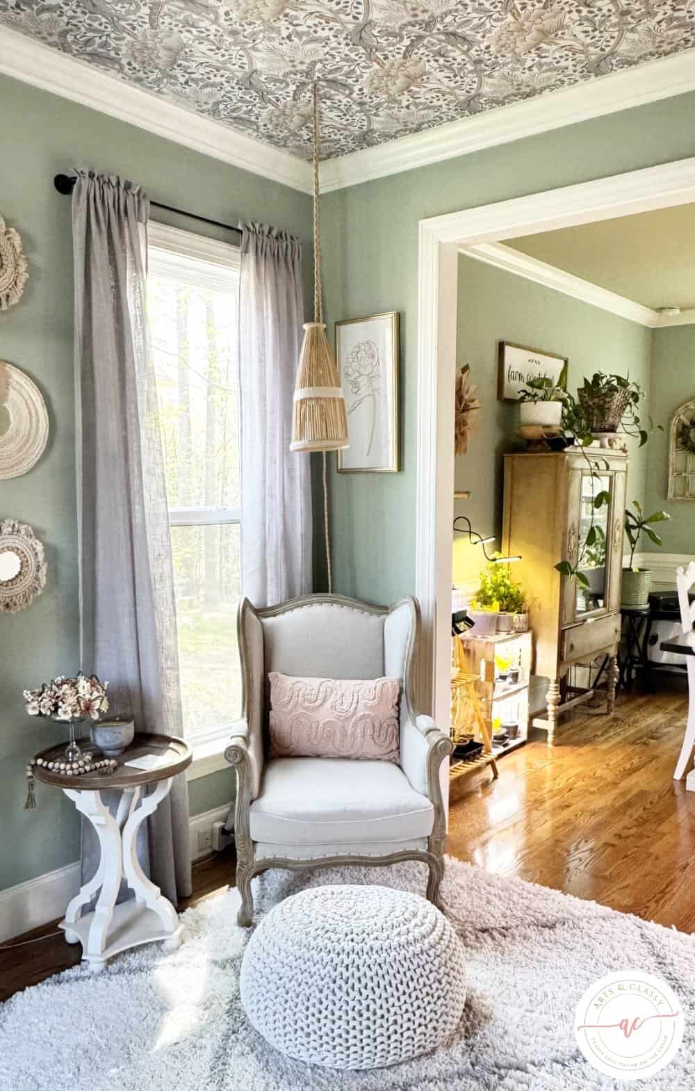 A cozy living room corner with an armchair, round knitted pouf, side table, woven pendant lamp, and patterned ceiling; sunlight streams through sheer curtains.