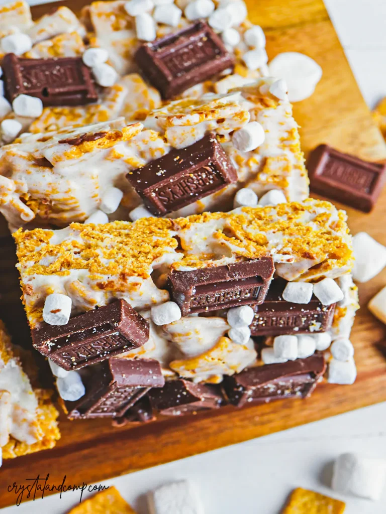 Close-up of s'mores dessert bars topped with mini marshmallows and pieces of chocolate on a wooden surface.
