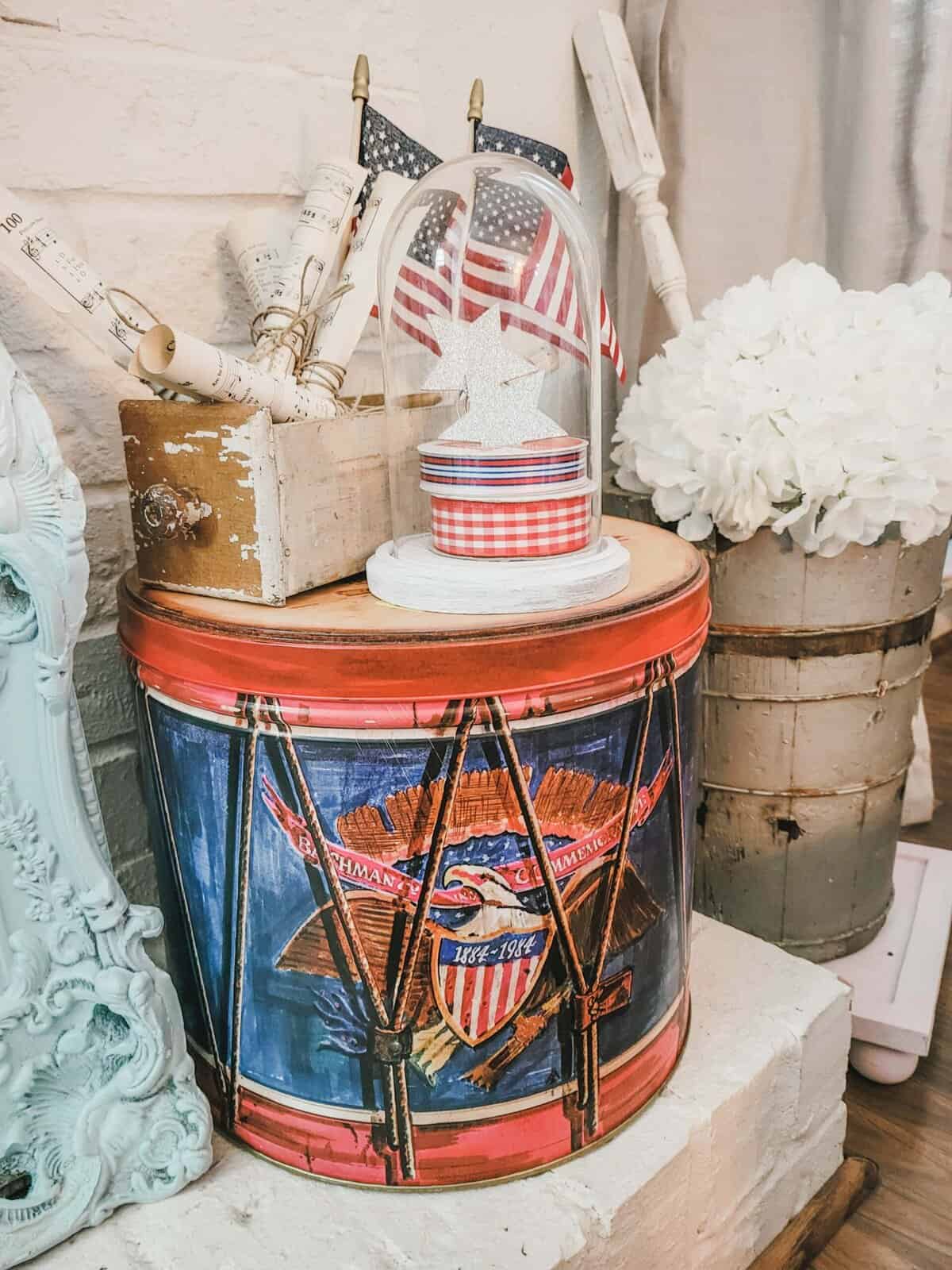 A decorative tin drum with American flags and a star sits on a white brick hearth, next to a white flower arrangement and rolled sheet music in a rustic container.