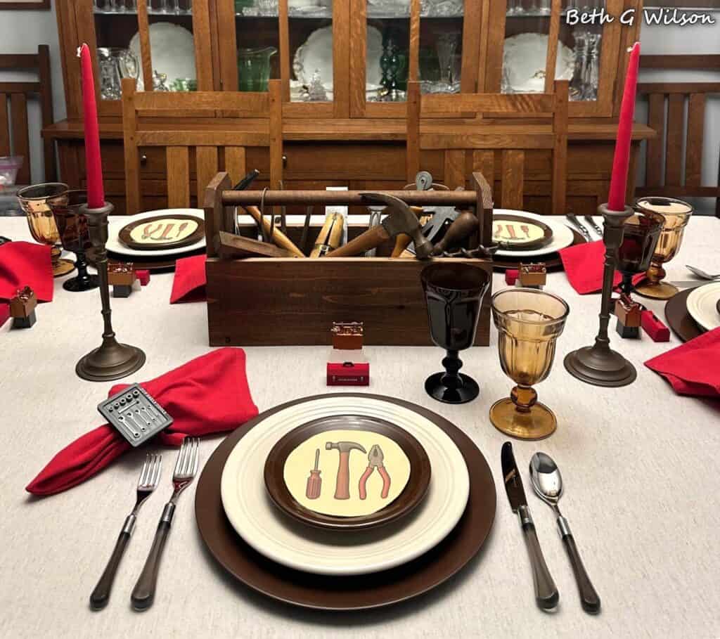 A dining table set with plates featuring tool illustrations, red candles, amber glasses, red napkins, and a wooden toolbox with tools as the centerpiece.