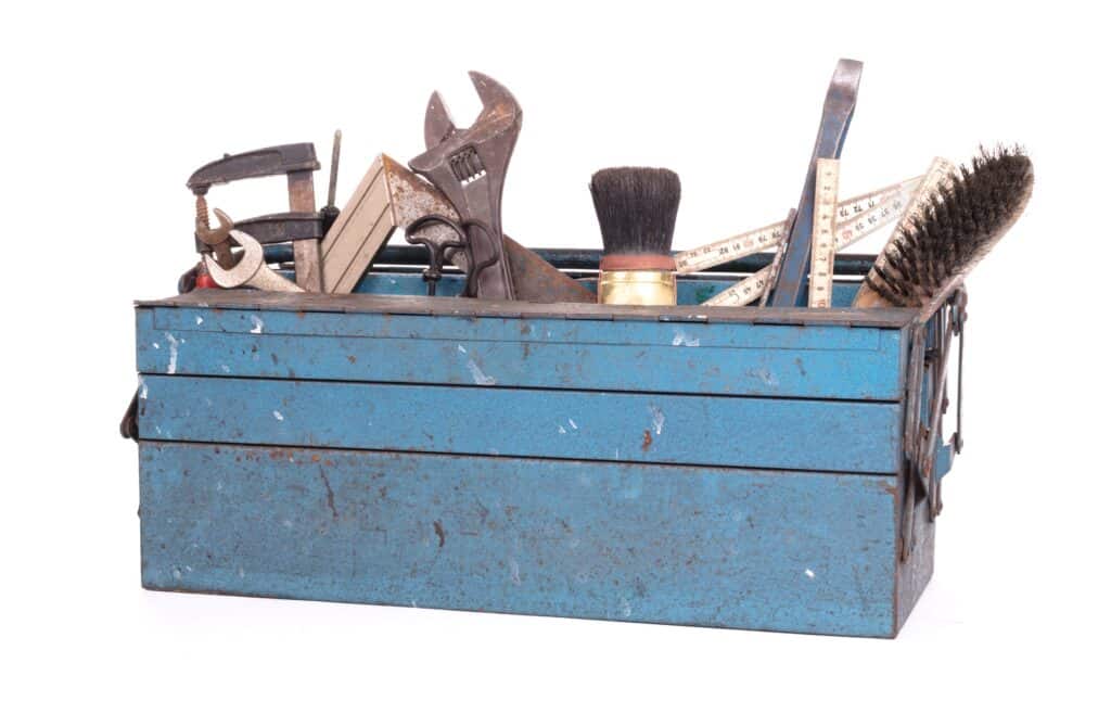 A blue metal toolbox containing assorted tools including a clamp, wrench, saw, paintbrush, ruler, and wire brush against a white background.