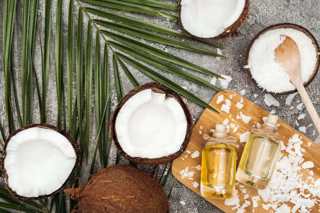 Coconut halves, shredded coconut, palm leaves, two small bottles of oil, and a wooden spoon are arranged on a gray surface.