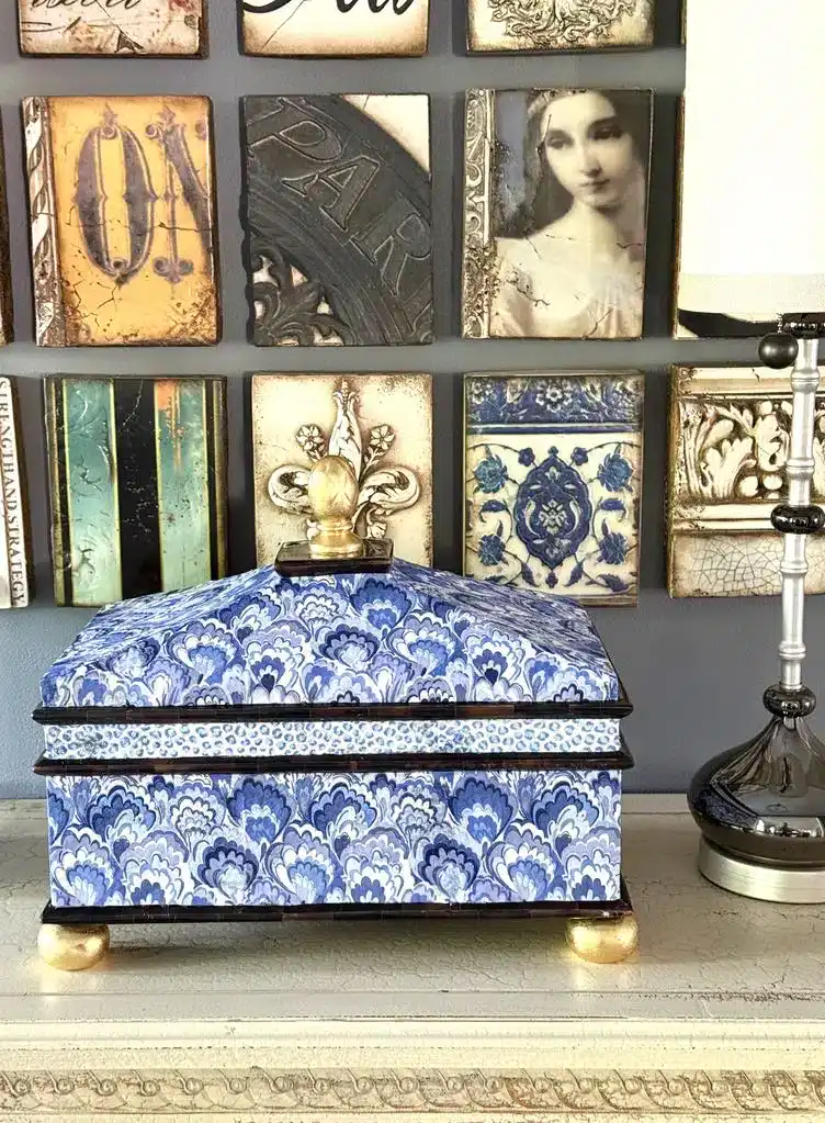 A blue and white patterned decorative box with gold feet and finial sits on a mantel, with a collage of framed art and a lamp in the background.