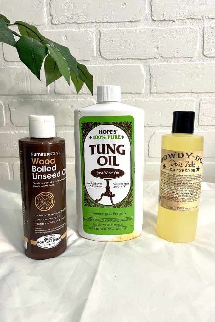 Three bottles of wood finishes—Wood Boiled Linseed Oil, Tung Oil, and Hemp Seed Oil—are displayed on a white surface against a white brick wall with a green leaf in view.