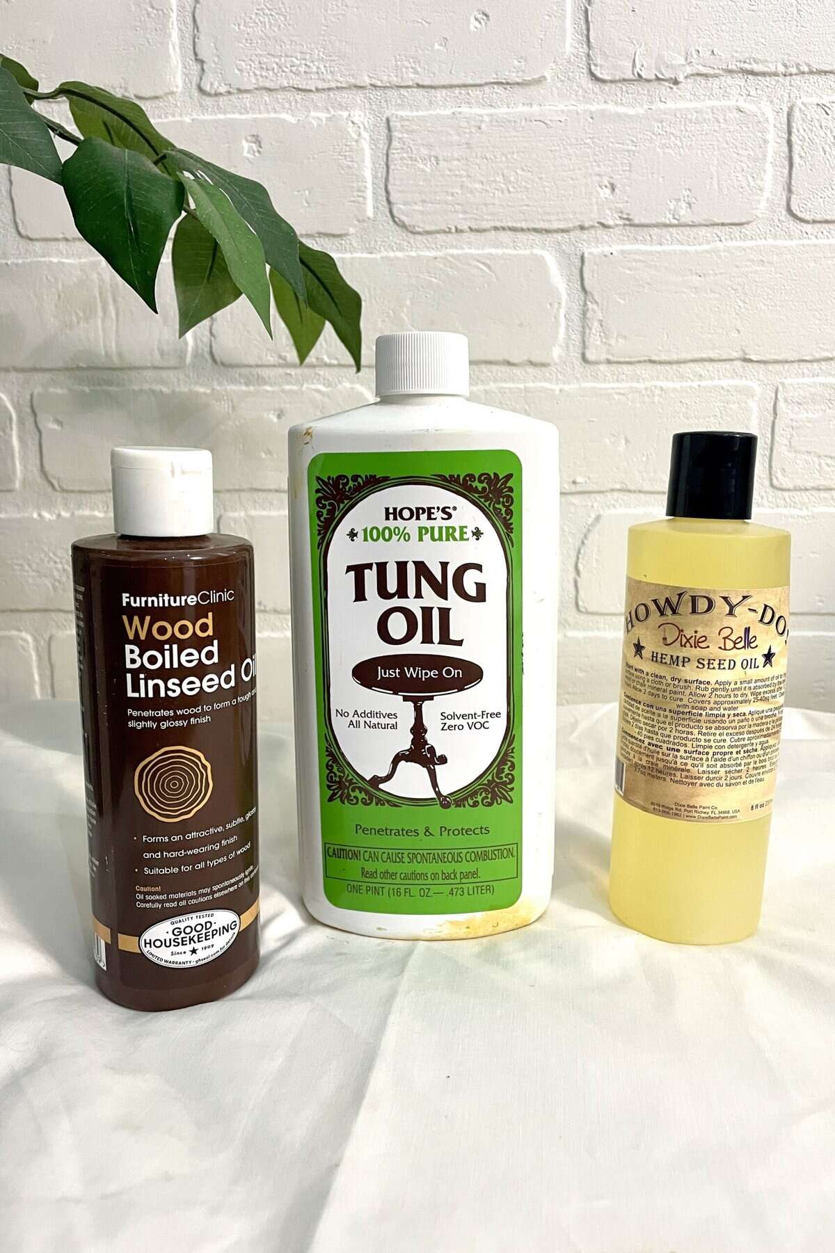 Three bottles of wood finishes—Wood Boiled Linseed Oil, Tung Oil, and Hemp Seed Oil—are displayed on a white surface against a white brick wall with a green leaf in view.