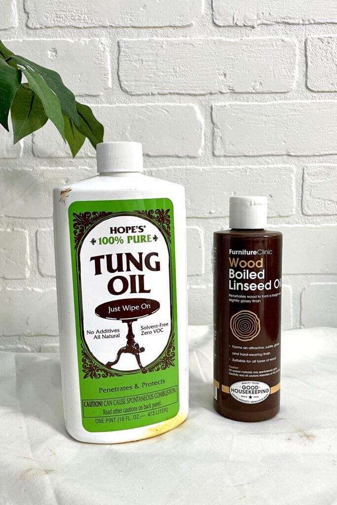 A bottle of Hope’s 100% Pure Tung Oil and a bottle of FurnitureClinic Wood Boiled Linseed Oil are placed side by side on a white surface in front of a white brick wall.
