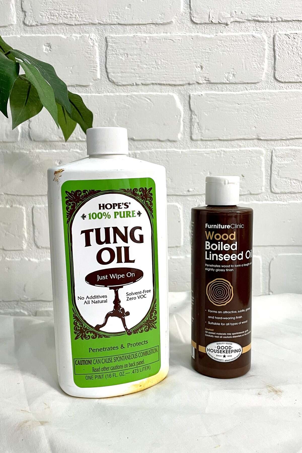 A bottle of Hope’s 100% Pure Tung Oil and a bottle of FurnitureClinic Wood Boiled Linseed Oil are placed side by side on a white surface in front of a white brick wall.