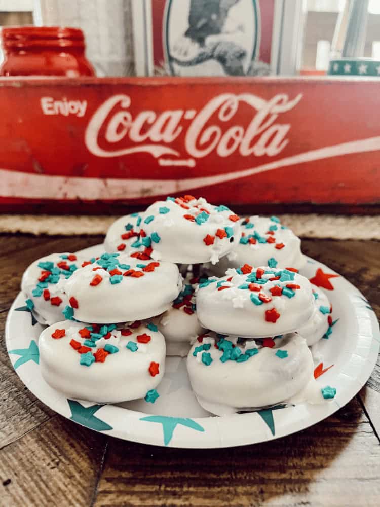 A paper plate holds white-frosted donuts topped with red, white, and blue star sprinkles. A vintage Coca-Cola sign is in the background.
