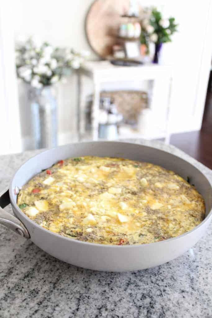 A cooked frittata with vegetables and cheese sits in a gray skillet on a granite countertop, with home decor in the blurred background.