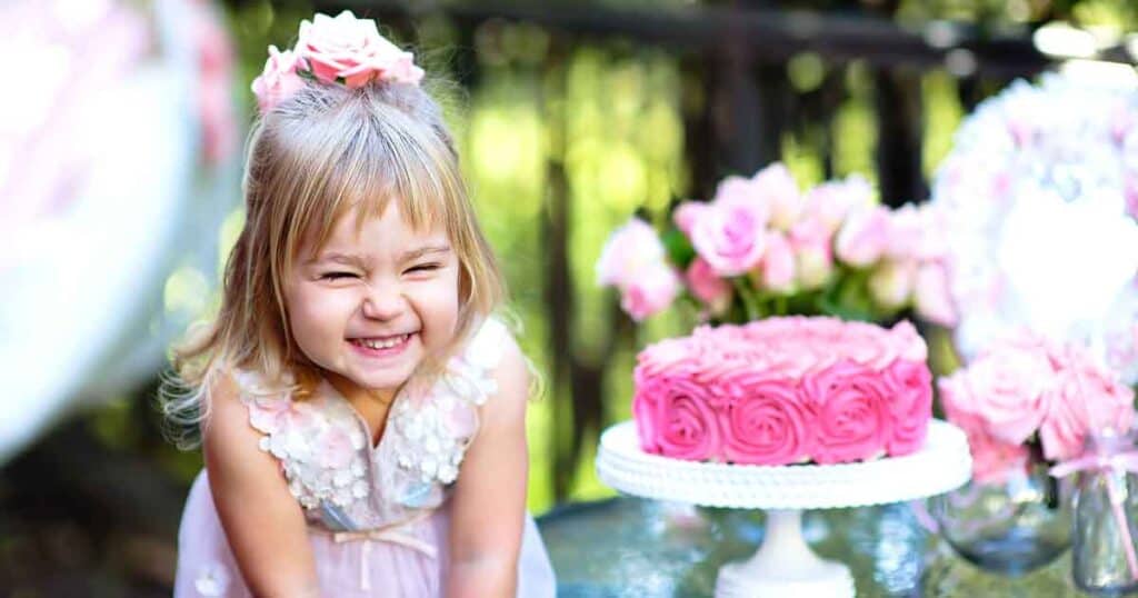 Young girl in a pink dress smiling in front of a pink rose-patterned cake on a stand, with pink flowers and greenery in the background.