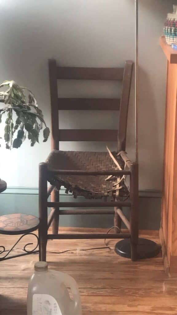 A wooden chair with a broken woven seat is positioned against a wall next to a wilted plant and a gallon water jug on the floor.