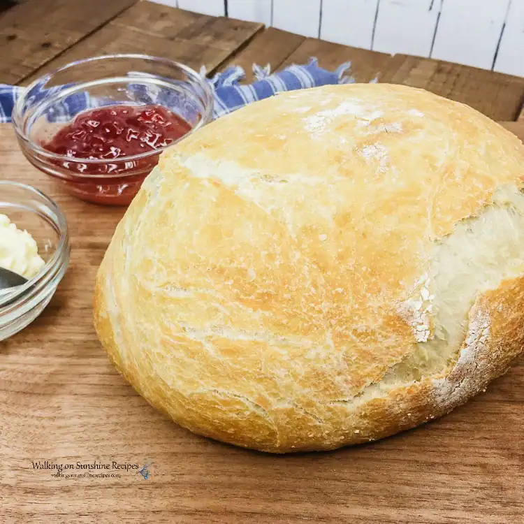 A round loaf of rustic bread on a wooden board with small bowls of butter and strawberry jam nearby.