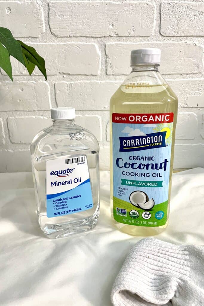 A bottle of mineral oil and a bottle of organic coconut cooking oil are placed side by side on a white surface with a washcloth nearby.