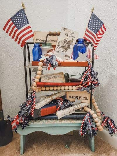 A small shelf decorated with American flags, patriotic signs, blue vases, rolled papers, and red, white, and blue garlands, displayed in a corner on a light blue bench.