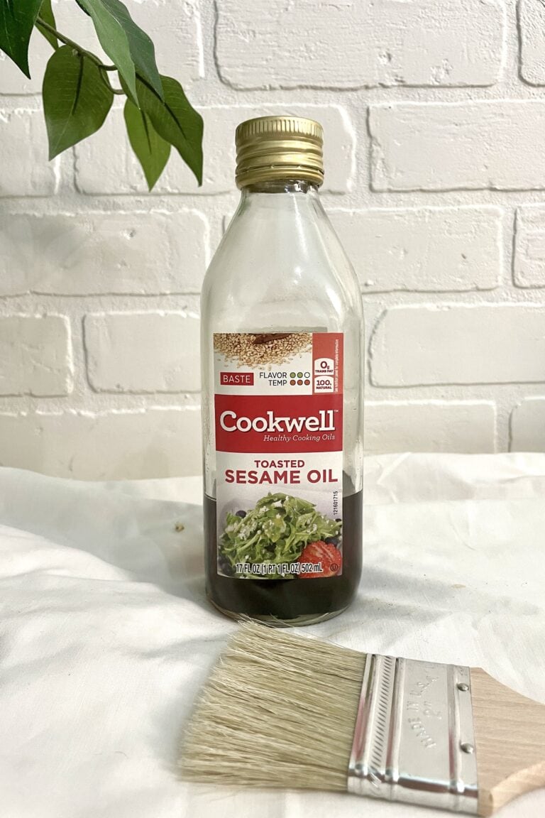 A bottle of Cookwell toasted sesame oil stands on a white surface next to a paintbrush, with a leafy plant and white brick wall in the background.