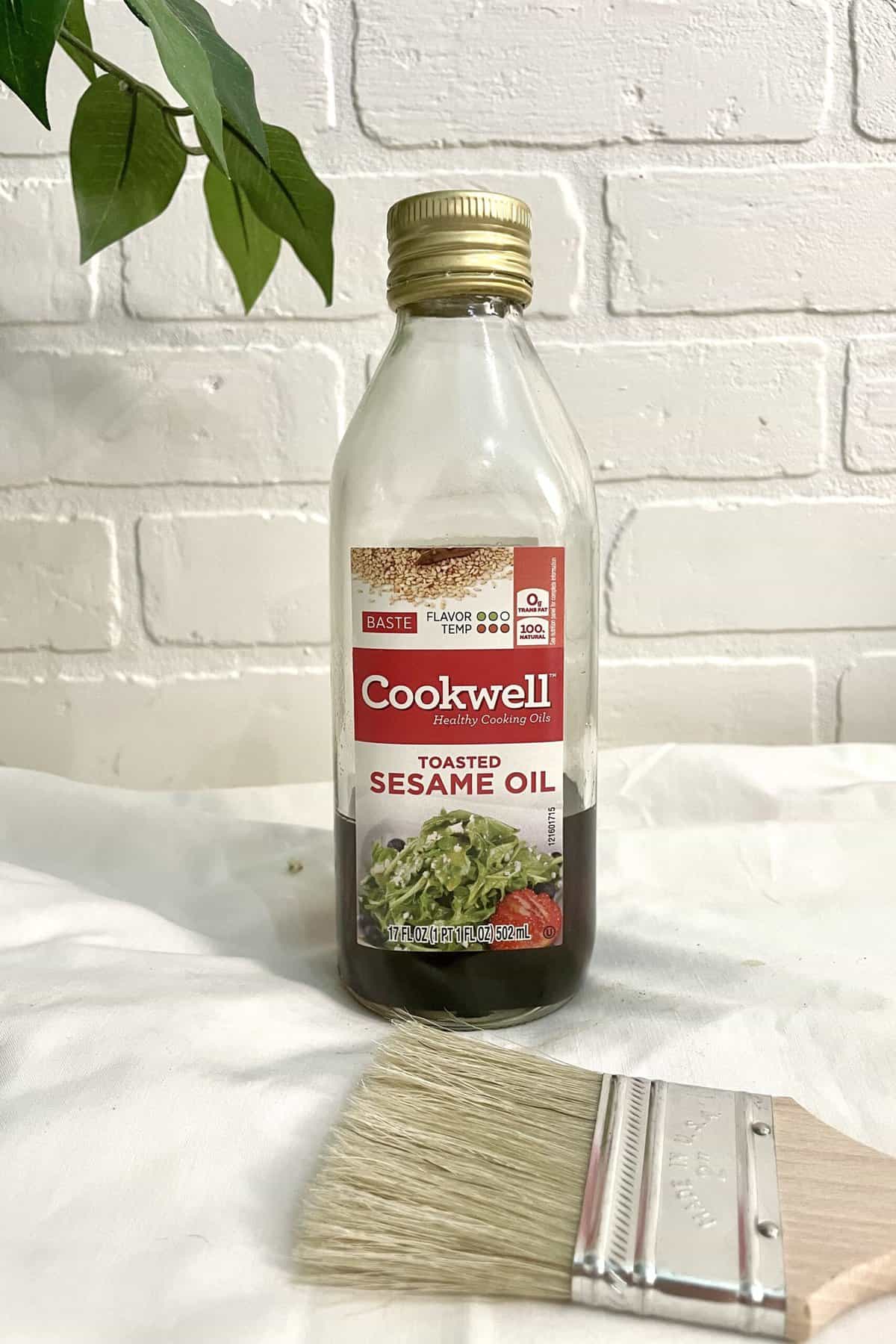 A bottle of Cookwell toasted sesame oil stands on a white surface next to a paintbrush, with a leafy plant and white brick wall in the background.