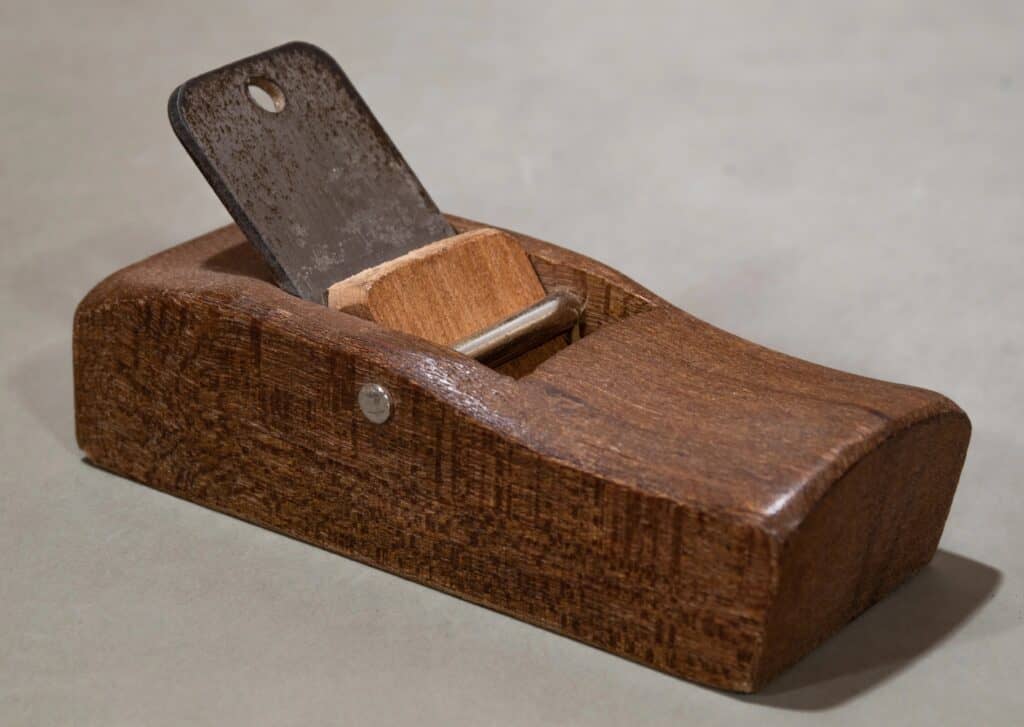 A wooden hand plane with a metal blade, used for smoothing or shaping wood surfaces, placed on a light gray surface.