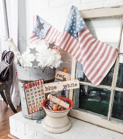 A rustic decor setup with a metal bucket holding white flowers, vintage bingo cards, red, white, blue tags, sewing items, and two American flag stockings hanging on a brick wall.