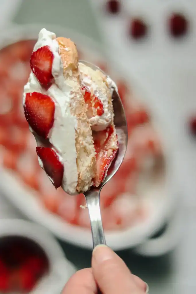 A spoon holds a layered dessert with sliced strawberries, whipped cream, and cake, with more of the same dessert blurred in the background.