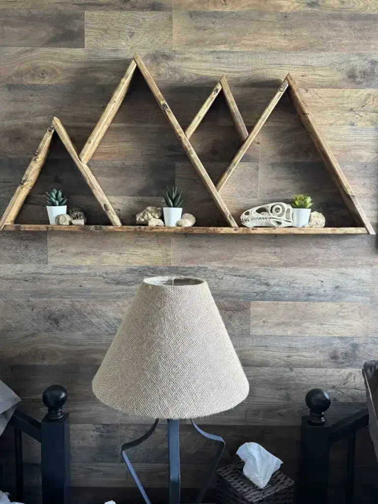 Wooden shelves shaped like mountain peaks display small potted plants and decorative items on a wood-paneled wall above a lamp with a burlap shade.