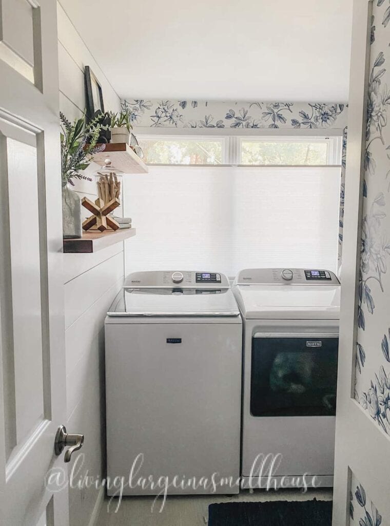 Laundry room with a washing machine and dryer side by side, shelves with decor above, floral wallpaper, and windows with white blinds.