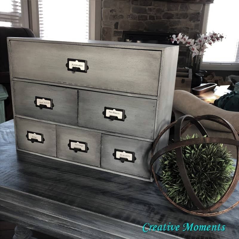 A small, vintage-style six-drawer cabinet with labeled metal handles sits on a wooden table next to a decorative orb with greenery, in a living room setting.