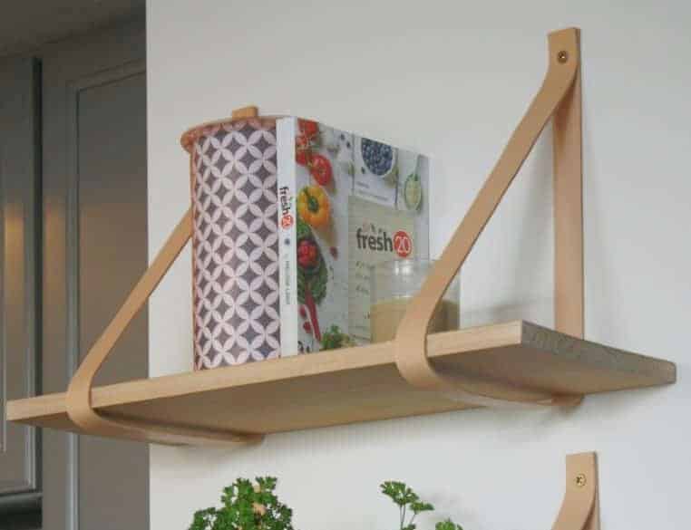 A wooden wall shelf with tan leather straps holds a patterned tin can, a cookbook titled "Fresh 20," and a small glass candle.