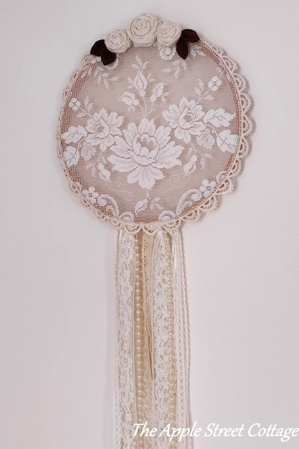 A round lace decor piece with floral embroidery, bordered with lace trim, topped with three fabric roses, and long lace ribbons hanging below.