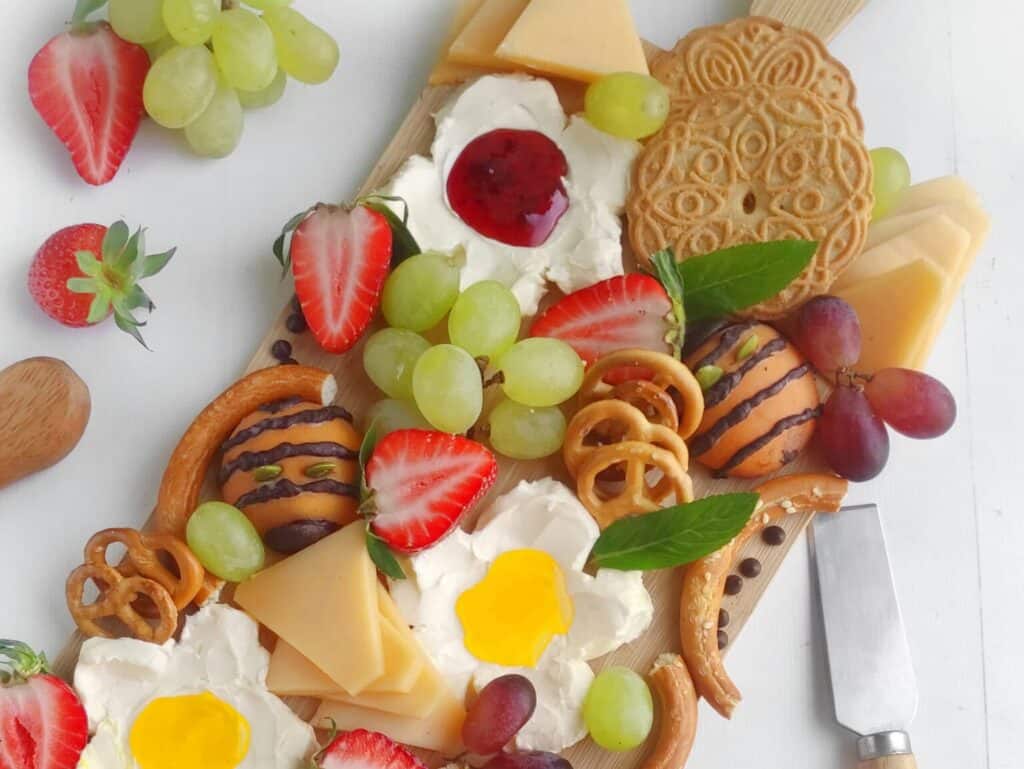 Charcuterie board with crackers, cheese slices, grapes, strawberries, pretzels, cookies, and cream cheese topped with jelly and egg-shaped decorations. A knife is placed nearby.