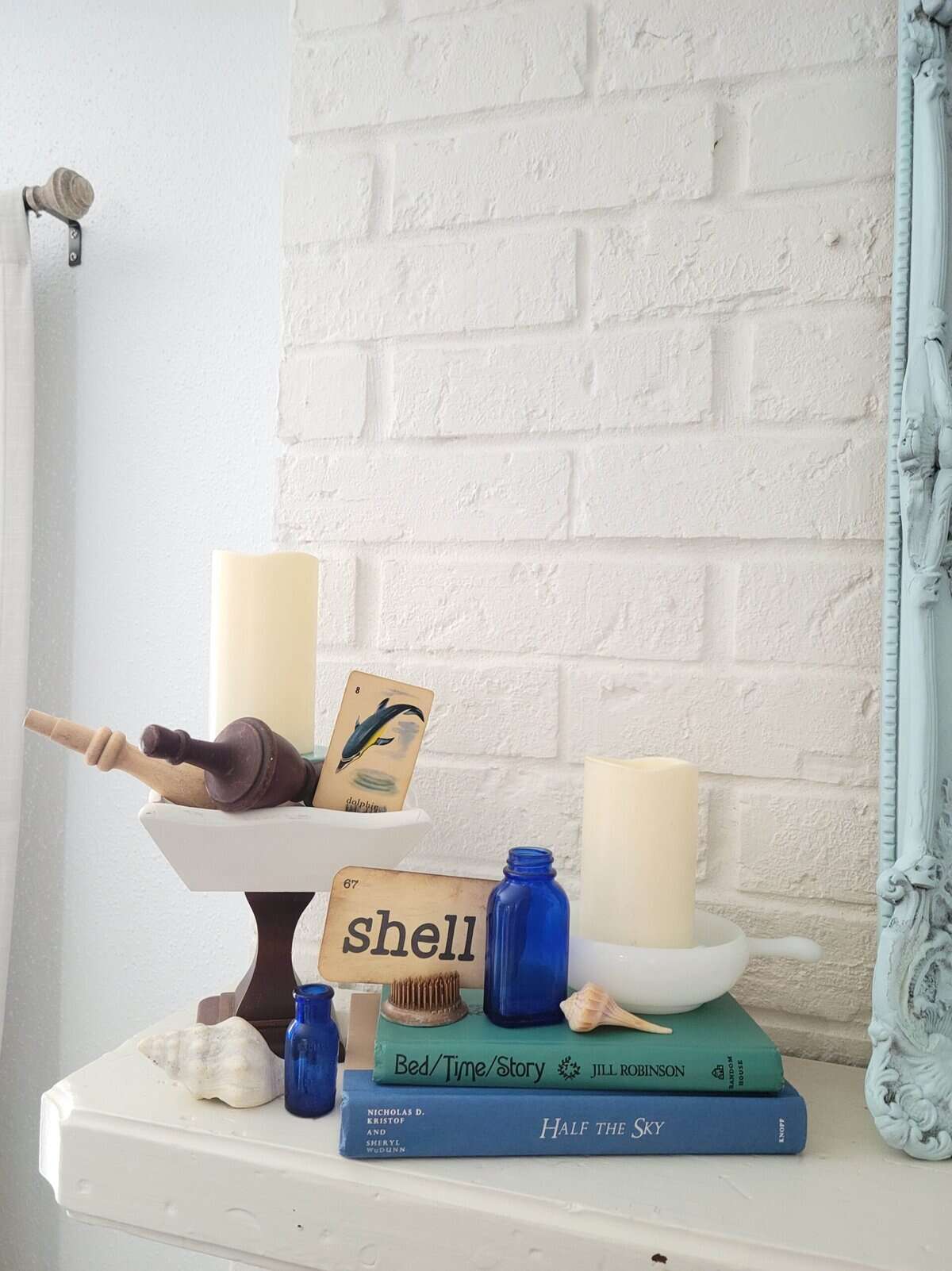 A white mantle displays candles, seashells, blue glass bottles, books, a wooden honey dipper, a playing card, and a tile with the word "shell" against a white brick wall.
