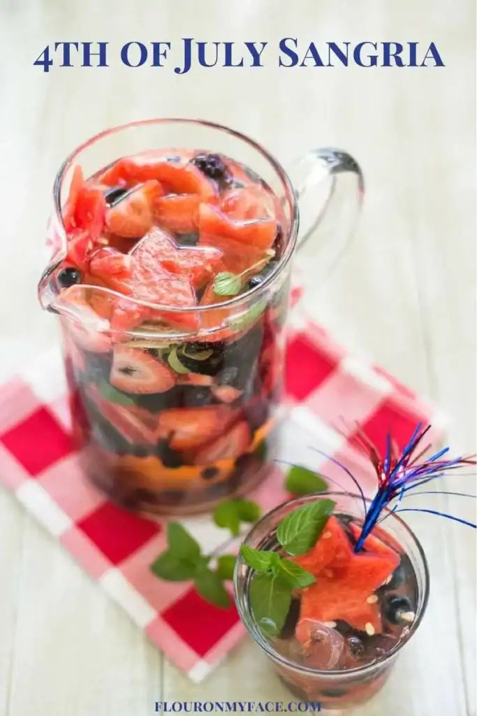 A pitcher and glass of sangria with watermelon, blueberries, and herbs sit on a red-and-white checkered napkin; text reads "4th of July Sangria.