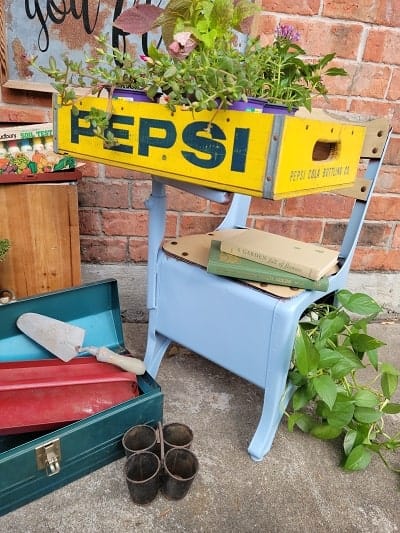 A yellow vintage Pepsi crate filled with plants sits on a light blue chair, surrounded by gardening tools and supplies against a brick wall.