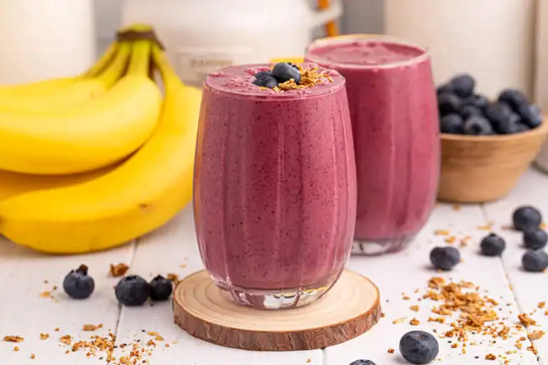 Two glasses of blueberry banana smoothie topped with granola and blueberries, with bananas and a bowl of blueberries in the background on a white surface.