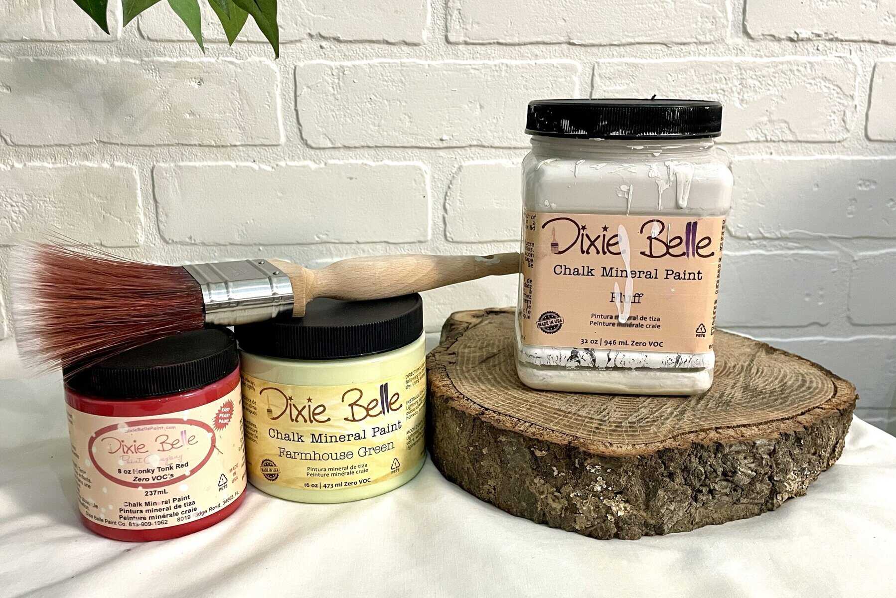 Three containers of Dixie Belle chalk mineral paint in red, yellow, and white, a paintbrush, and a wood slice are displayed against a white brick wall background.
